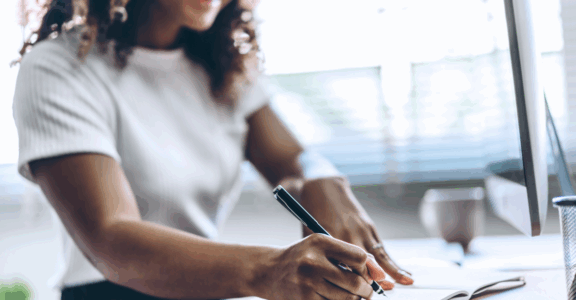 A person writes in a notebook at a computer desk